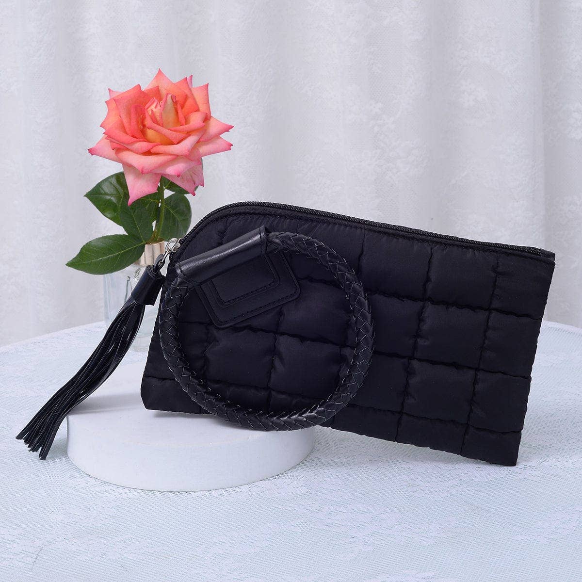 Black quilted clutch bag with circular handle and tassel, placed on a white surface with a pink rose in the background.
