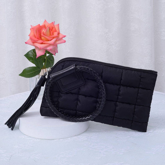 Black quilted clutch bag with circular handle and tassel, placed on a white surface with a pink rose in the background.