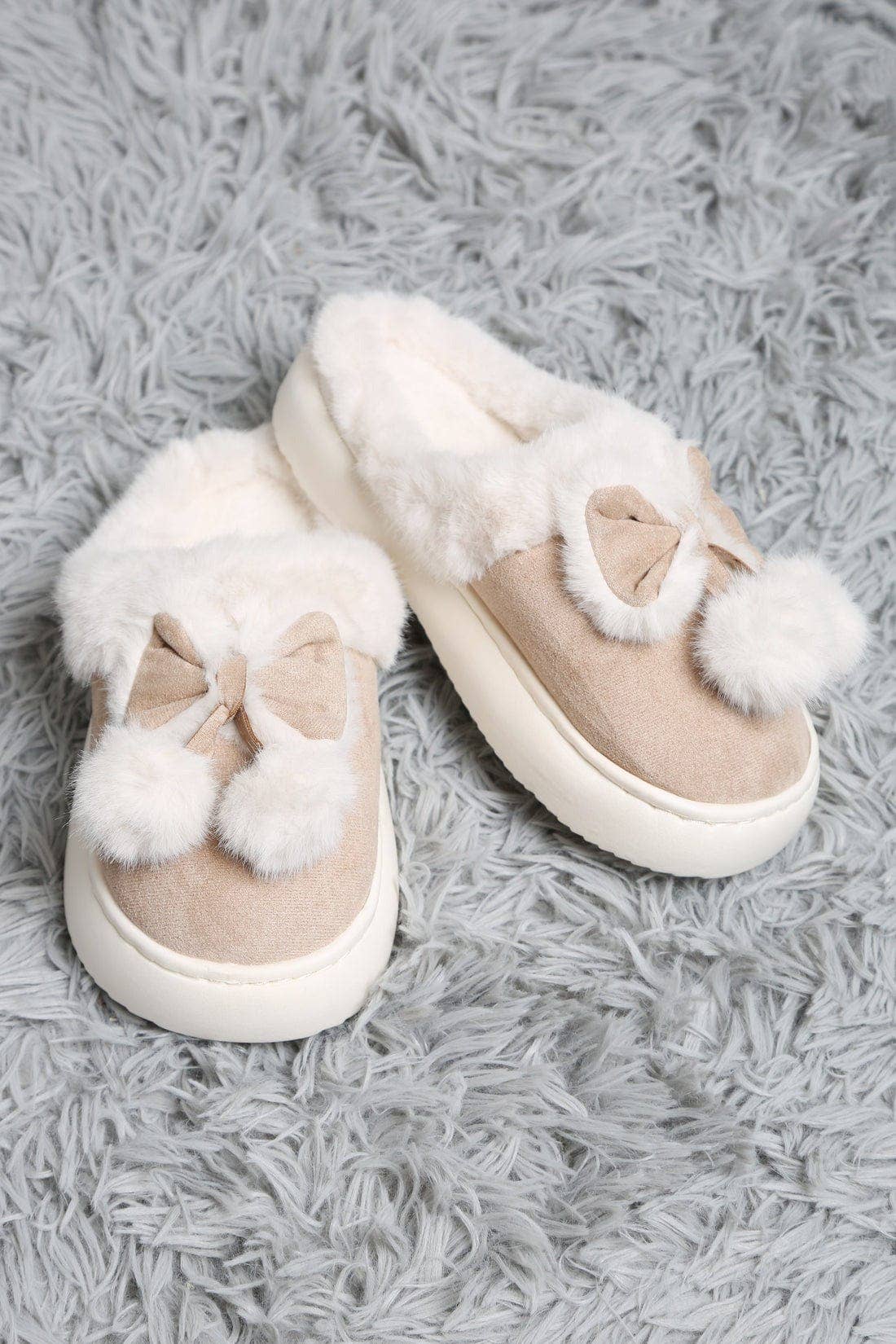 Pair of beige slippers with white fur pom-poms on a gray textured surface